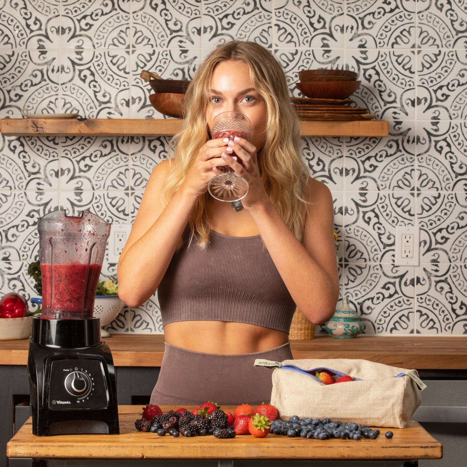 Woman drinking a fresh berry smoothie in a kitchen with reusable linen produce bags filled with fruit.